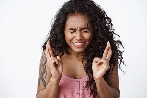 Primer plano mujer de pelo rizado ansiosa y preocupada con tatuajes, entrecerrar los ojos, apretar los dientes nerviosamente, cruzar los dedos, buena suerte, anticipar algo importante, orar de pie pared blanca