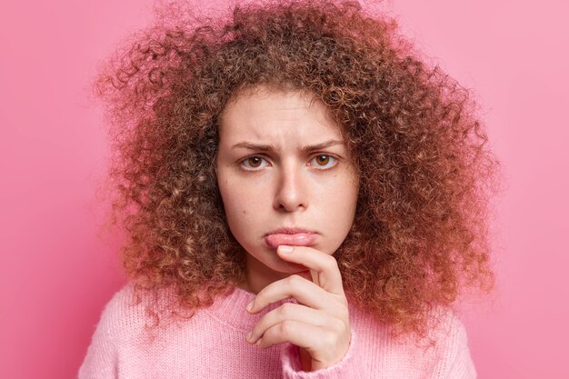 Primer plano de una mujer de pelo rizado disgustada que frunce los labios, se siente triste, frunce el ceño, tiene expresión de descontento y usa un jersey casual aislado sobre una pared rosa. Concepto de expresiones faciales negativas