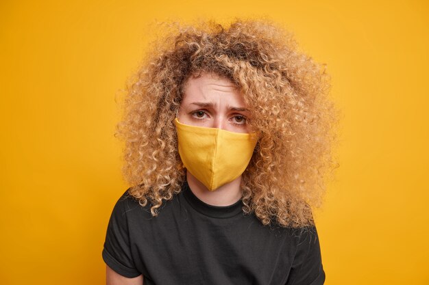 Primer plano de una mujer de pelo rizado que se ve tristemente lleva una máscara protectora harta de las restricciones de encierro viste una camiseta negra aislada sobre una pared amarilla. Pandemia de coronavirus