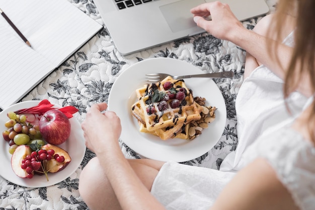 Primer plano de una mujer con plato de waffle y frutas usando laptop