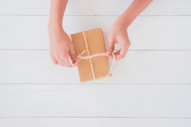 Foto gratuita primer plano de una mujer que ata el hilo en una caja de regalo envuelta en marrón en el escritorio de madera