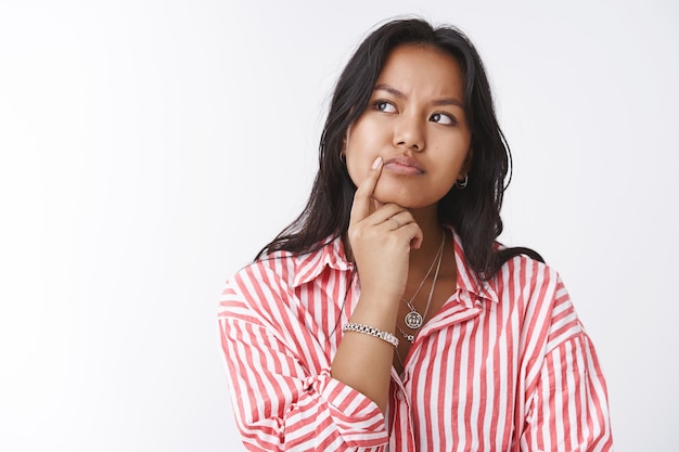 Primer plano de una mujer reflexiva y enfocada que tiene un pensamiento problemático difícil, toma una decisión en mente, frunciendo el ceño, tocando el labio y mirando la esquina superior izquierda, pensando contra el fondo blanco.