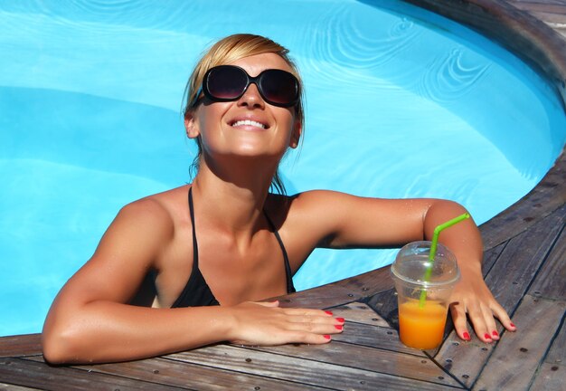 Primer plano de una mujer rubia sonriente con una taza de jugo en la piscina