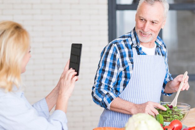 Primer plano de una mujer senior tomando una foto de su esposo preparando la ensalada en el tazón en el teléfono móvil