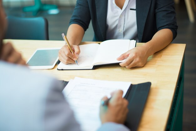 Primer plano de mujer sentada a la mesa y escribiendo notas en el bloc de notas