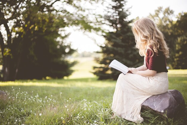 Primer plano de una mujer sentada sobre una roca y leyendo la Biblia en un campo de hierba