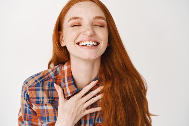 Primer plano de mujer sonriente feliz con cabello rojo natural y piel pálida, sosteniendo la mano en el corazón y riendo con los ojos cerrados