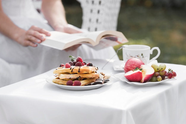 Primer plano de una mujer sosteniendo un libro en la mano sentado detrás de la mesa de desayuno al aire libre