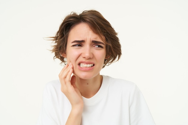 El primer plano de una mujer tiene dolor de muelas, se toca los dientes y frunce el ceño debido a una dolorosa incomodidad.