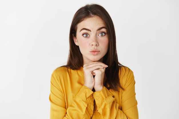 Primer plano de una mujer tonta intrigada y emocionada mirando al frente con interés, mirándote como si estuvieras parado con una camisa amarilla sobre una pared blanca