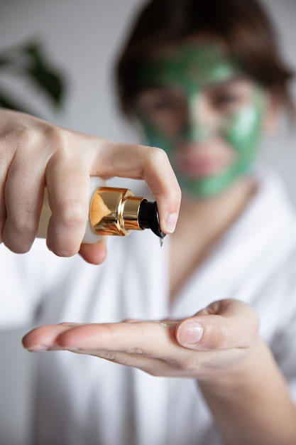 Primer plano, una mujer con una túnica blanca con una máscara cosmética verde presiona sobre el dispensador de productos para el cuidado de la belleza, fondo borroso.