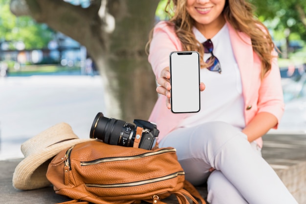 Primer plano de mujer turista sentada al lado de la bolsa; sombrero y cámara mostrando su pantalla de teléfono móvil