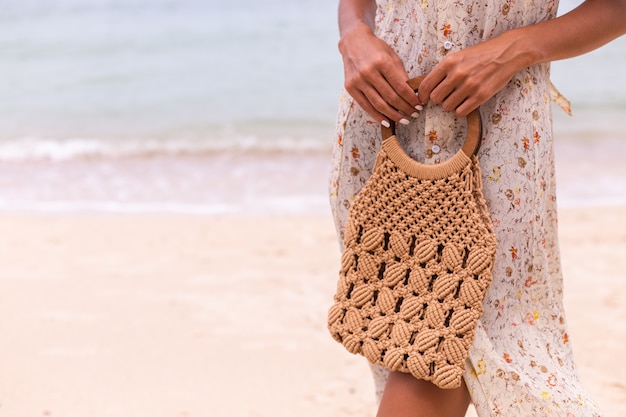 Primer plano de mujer en vestido de vuelo de verano ligero con bolso tejido en la playa, mar de fondo.