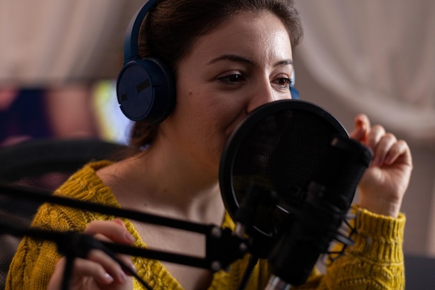 Primer plano de una mujer vlogger con auriculares hablando por micrófono de producción preparando podcast en línea en casa en la sala de estar. Blogger influencer transmitiendo contenido en vivo respondiendo a una pregunta