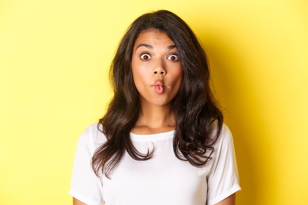 Primer plano de una niña afroamericana tonta y divertida, chupando los labios y mirando sorprendido, de pie contra el fondo amarillo