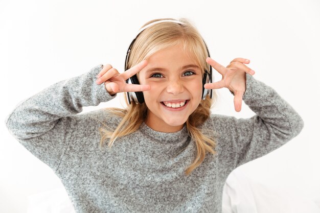 Primer plano de niña divertida en hedphones mostrando gesto de paz con las dos manos, mirando a la cámara