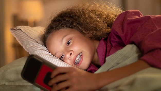 Primer plano de una niña feliz acostada en la cama con el teléfono. Linda chica rizada vestida con pijamas rosas jugando al teléfono, alegre y sonriente viendo la pantalla. Familia, niños descansan y usan el concepto de dispositivos inteligentes