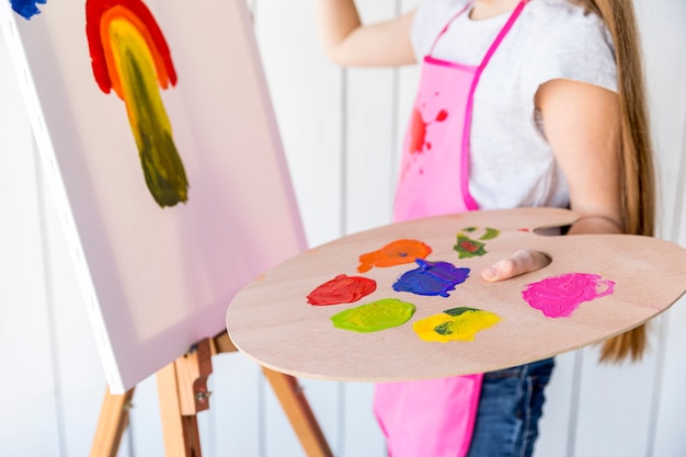 Primer plano de una niña pintando sobre lienzo con paleta de madera multicolor en la mano