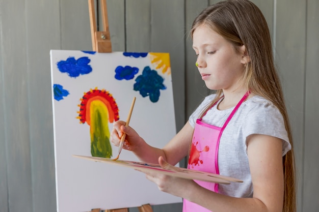 Primer plano de una niña que mezcla la pintura con un cepillo de pie delante de un lienzo pintado