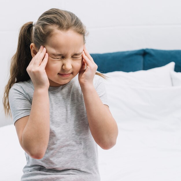 Primer plano de una niña sentada en la cama que tiene dolor severo en dolor de cabeza