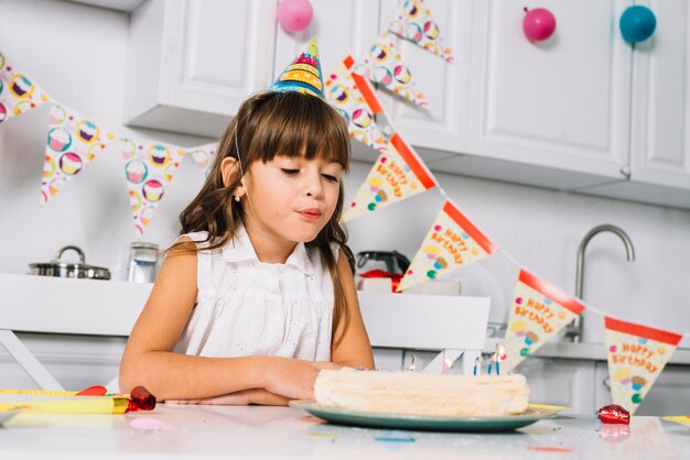 Primer plano de una niña con sombrero de fiesta en la cabeza soplando velas en el pastel