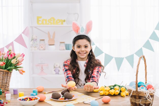 Primer plano de una niña sonriente con orejas de conejo mostrando coloridos huevos de Pascua
