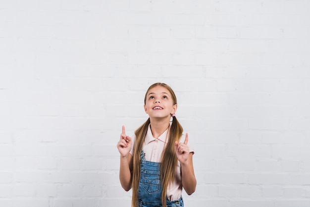 Foto gratuita primer plano de una niña sonriente que señala el dedo hacia arriba mirando hacia arriba de pie contra la pared de ladrillo blanco