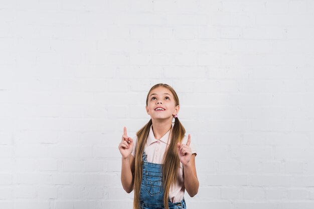 Primer plano de una niña sonriente que señala el dedo hacia arriba mirando hacia arriba de pie contra la pared de ladrillo blanco
