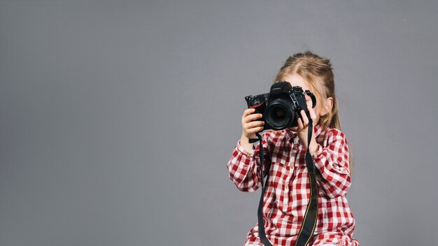 Primer plano de una niña sosteniendo una cámara frente a su cara de pie contra el fondo gris