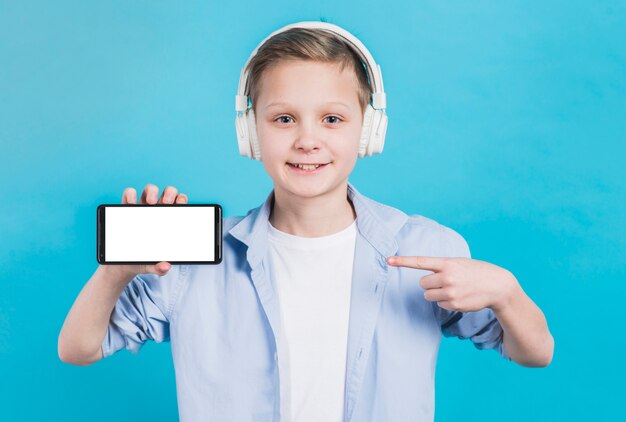 Primer plano de un niño con auriculares en la cabeza apuntando su dedo hacia el teléfono móvil con pantalla en blanco