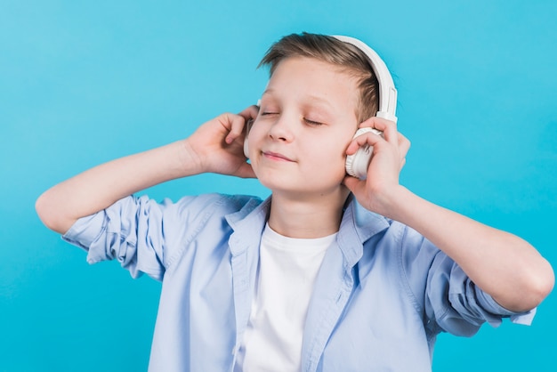 Primer plano de un niño disfrutando de la música que escucha en los auriculares blancos contra el fondo azul