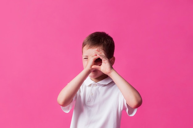 Primer plano del niño mirando a través de la mano como binoculares sobre fondo de pared rosa