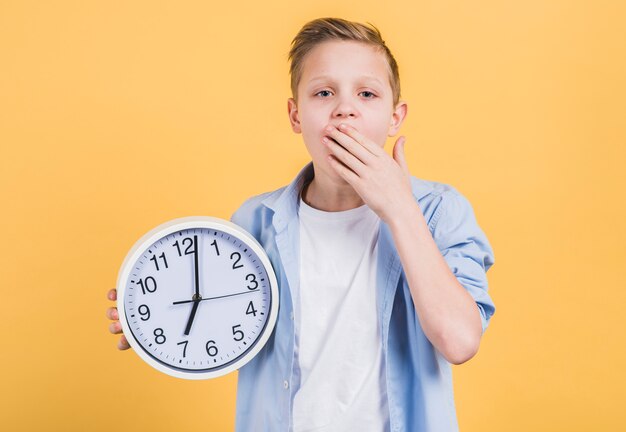 Primer plano de un niño que sostiene un reloj blanco redondo que bosteza con su mano en la boca de pie contra el fondo amarillo