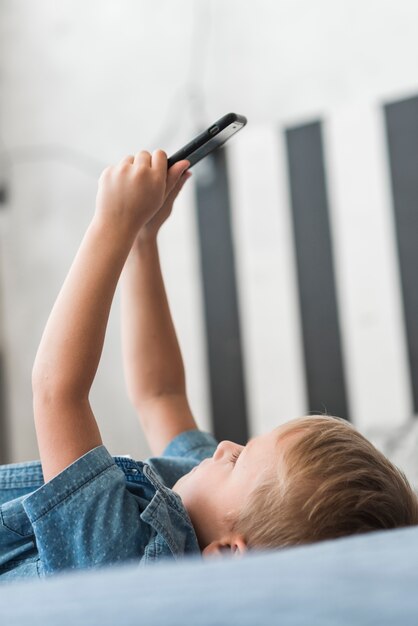 Primer plano de un niño rubio acostado en la cama usando un teléfono móvil