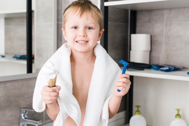Primer plano de un niño sonriente de pie en el baño con brocha de afeitar y maquinilla de afeitar azul en la mano