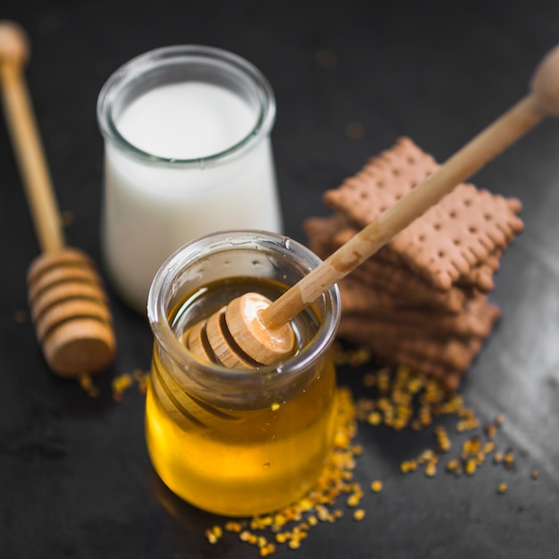 Primer plano de la olla de leche; tarro de miel; galletas y polen de abeja en el fondo negro