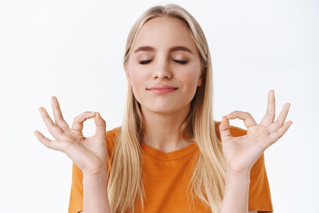 Primer plano, pacífica, decidida y con una mente sana, atractiva chica rubia con camiseta naranja, ojos cerrados haciendo gestos zen, meditando con los ojos cerrados y sonriendo aliviada, haciendo práctica de respiración de yoga