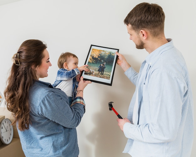 Primer plano de una pareja feliz con su bebé pequeño fijando marco en la pared