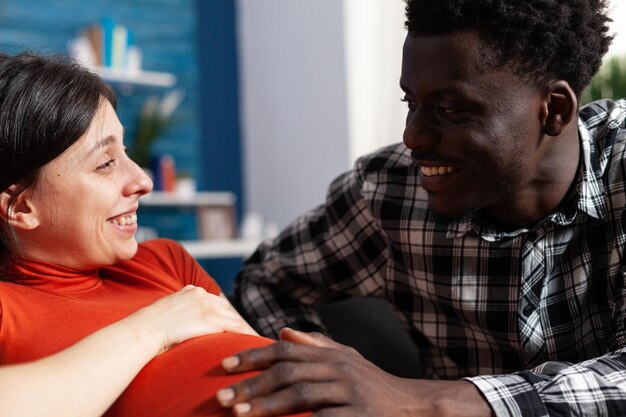 Primer plano de una pareja joven interracial sonriente esperando recién nacido mientras está sentado en casa. Parejas casadas relajadas enamoradas esperando a un niño por nacer mientras el marido afroamericano se siente caucásico embarazada