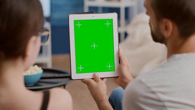 Primer plano de una pareja sosteniendo una tableta digital vertical con pantalla verde en una conferencia en línea o una videollamada grupal en la sala de estar de la casa. Hombre y mujer hablando frente a un dispositivo de pantalla táctil con llave de croma.