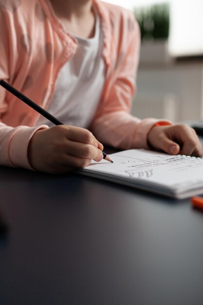Primer plano de un pequeño alumno escribiendo la tarea de matemáticas en el cuaderno durante la lección en el aula sentado en la mesa de escritorio en la sala de estar. Concepto de educación en el hogar, educación a distancia, educación en línea