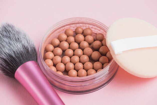 Primer plano de perlas bronceadoras; cepillo de esponja y maquillaje sobre fondo rosa