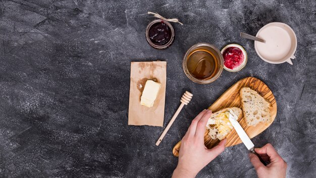 Primer plano de una persona agregando la mantequilla con un cuchillo; Mermelada de frambuesa y miel sobre fondo negro con textura