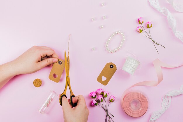 Primer plano de una persona cortando la etiqueta con una pulsera; Rosas artificiales y cinta sobre fondo rosa.