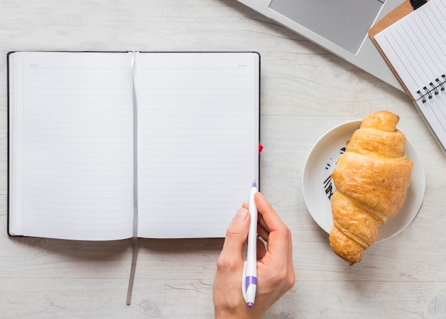 Primer plano de una persona que escribe en el diario con lápiz y croissant en un plato sobre el escritorio de madera