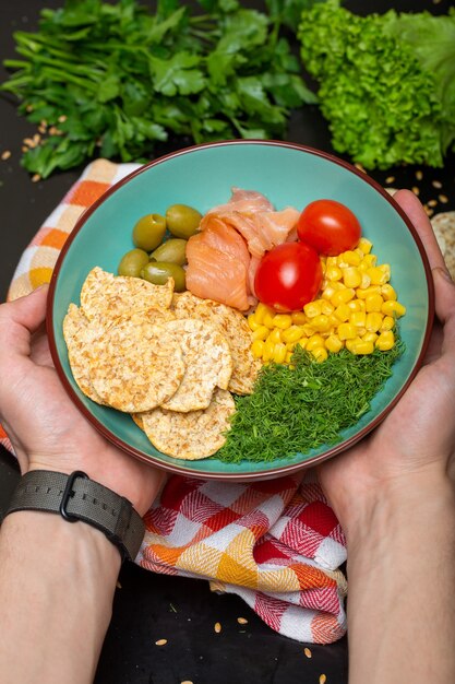 Primer plano de una persona que sostiene un plato de ensalada con salmón, galletas saladas y verduras bajo las luces