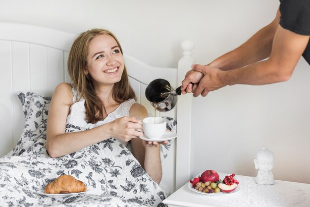 Primer plano de una persona que vierte una bebida caliente en la taza sostenida por una mujer sentada en la cama