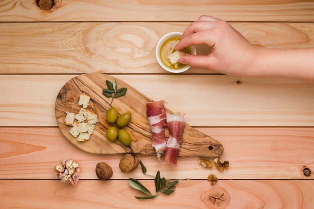 Primer plano de una persona sumergiendo la rebanada de pan en aceituna infundida con tocino; Oliva y nueces en mesa de madera.