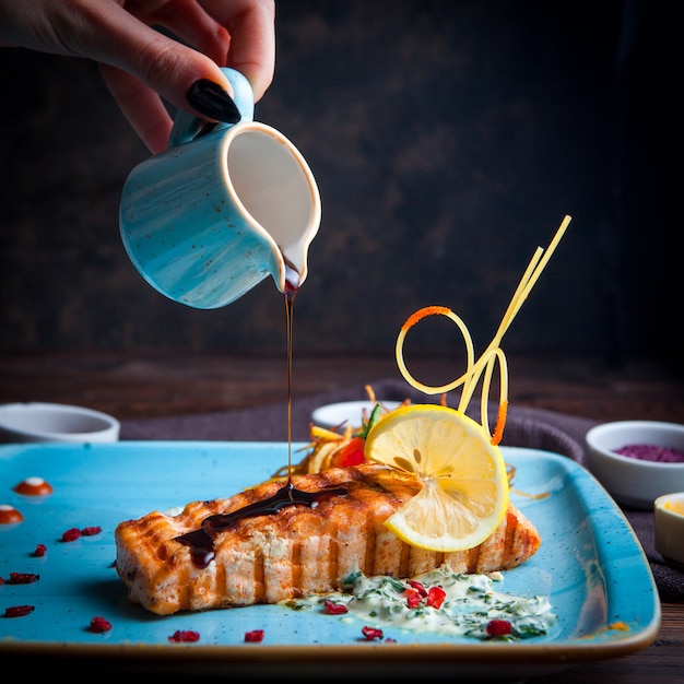 Primer plano de pescado a la parrilla, salsa vertiendo a mano con guarnición, limón, salsa y especias en un plato azul sobre una mesa de madera oscura