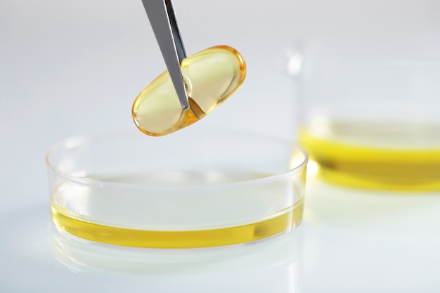 Primer plano de pinzas sosteniendo una cápsula de color amarillo claro sobre líquido amarillo en un plato de vidrio en un laboratorio